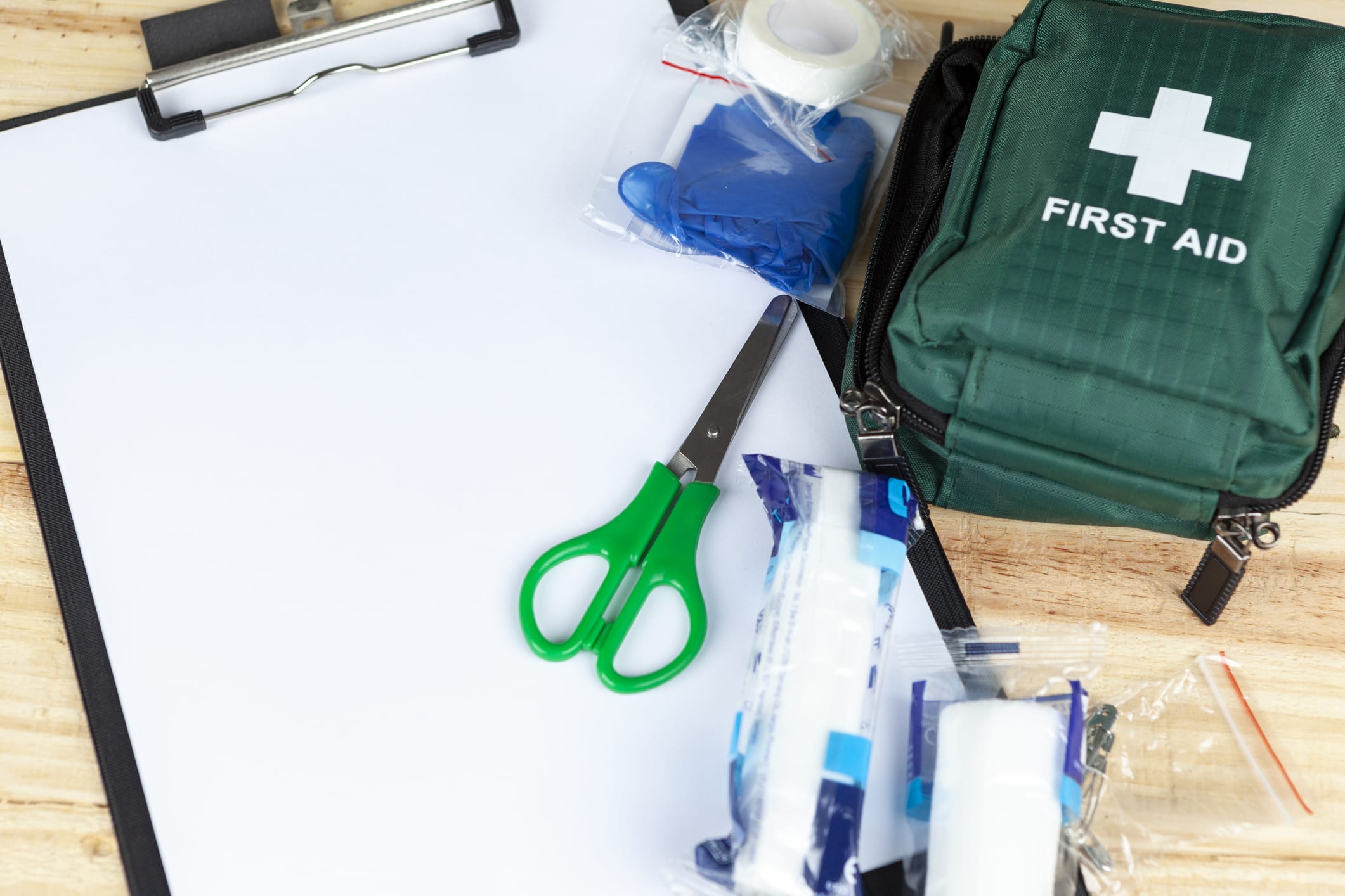 Green first aid kit on a wooden table with a clipboard and a pair of scissors and some dressings