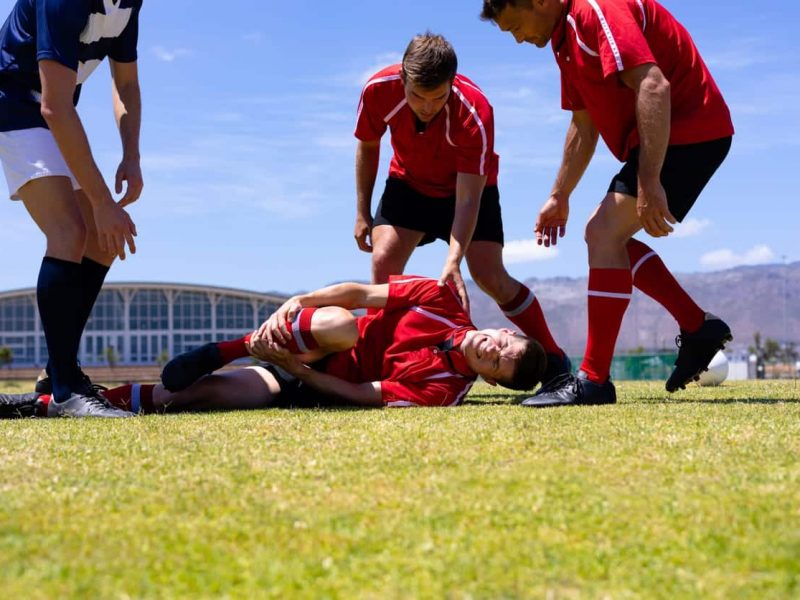 Low angle front view of a group of Caucasian male rugby players from opposing teams wearing team uniforms, gathered around an injured player lying the ground clutching his leg on a rugby pitch during a match, with blue sky in the background